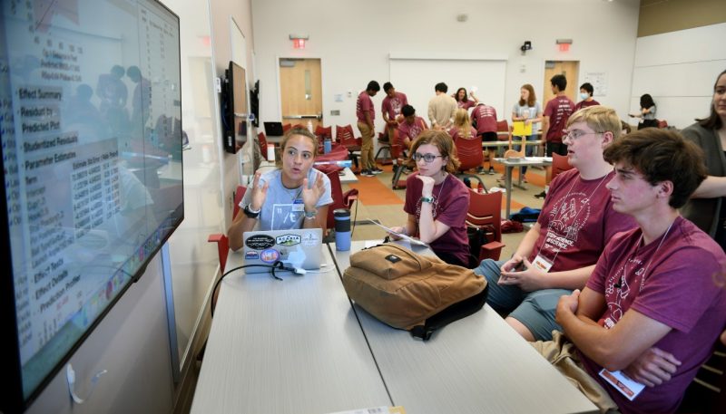 Students sit around a table discussing data results that are displayed on a large screen.